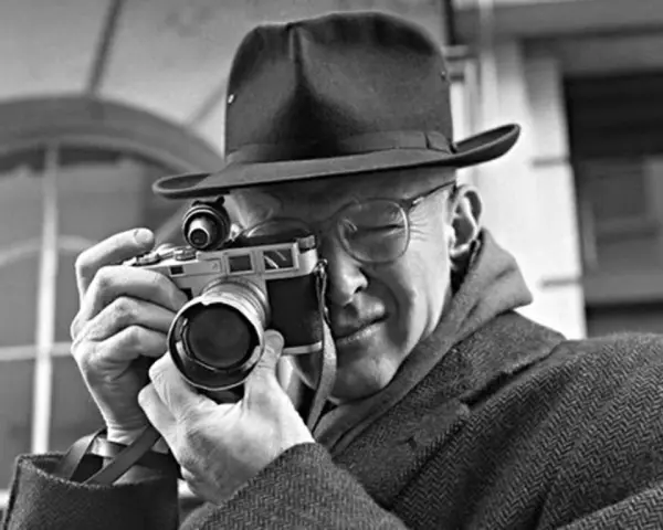 Mężczyzna w kapeluszu i okularach trzyma aparat, gotowy do zrobienia zdjęcia. Wśród znanych fotografów, jego postawa sugeruje skupienie i profesjonalizm.