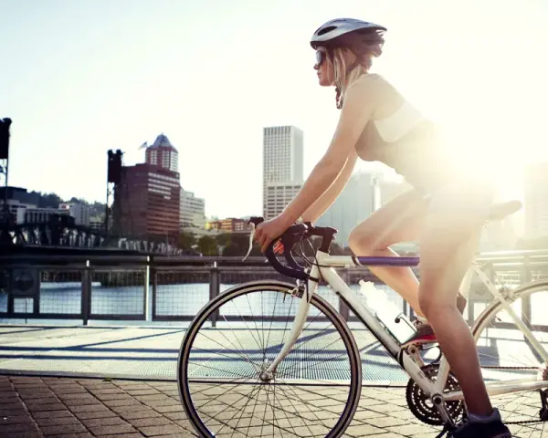 A female cyclist enjoys a sunny ride along a waterfront path with a city skyline in the background.