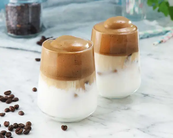 Två glas med krämigt dalgona kaffe med en fluffig kaffetopping över mjölk och is. Kaffebönor sprider sig på marmorbordet.