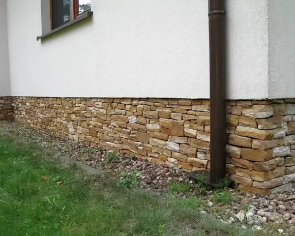 Eleganckie cokoliki z kamienia ozdabiają dom, tworząc naturalną, kamienną opaskę wokół białej ściany.