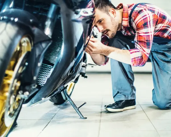 Mechanik w kratkę wykonuje przegląd motocykla, pochylając się nad jego przednią częścią.