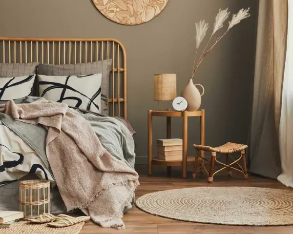 Dormitorio con cama de ratán, ropa de cama beige y gris, manta rosa, mesita auxiliar de madera y alfombra redonda.
