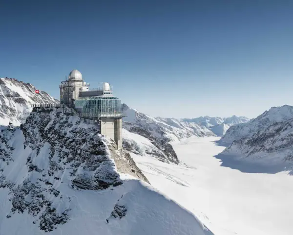 A modern building with glass walls and domes sits atop a snowy mountain, offering breathtaking views of the surrounding peaks and a vast glacier. This is the top of Europe, a place of incredible altitude.