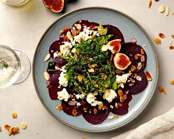 Rote Bete Carpaccio mit Feigen, Ziegenkäse und Mandelsplittern auf einem blauen Teller, daneben ein Glas Weißwein.