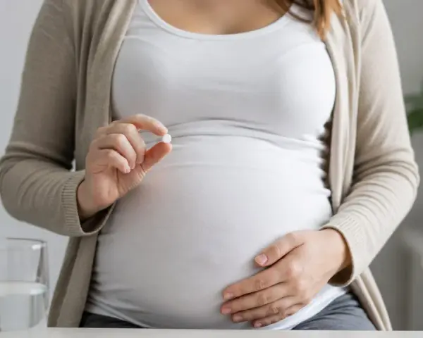 Kobieta w ciąży trzyma tabletkę, zastanawiając się nad aspiryną w ciąży. Obok stoi szklanka wody.