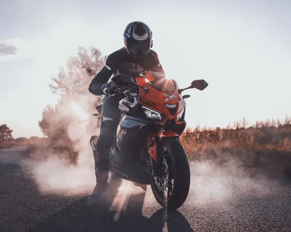 Motocyklista w kasku i kombinezonie na pomarańczowym motocyklu Barton Blade, z opon dymiących na drodze.