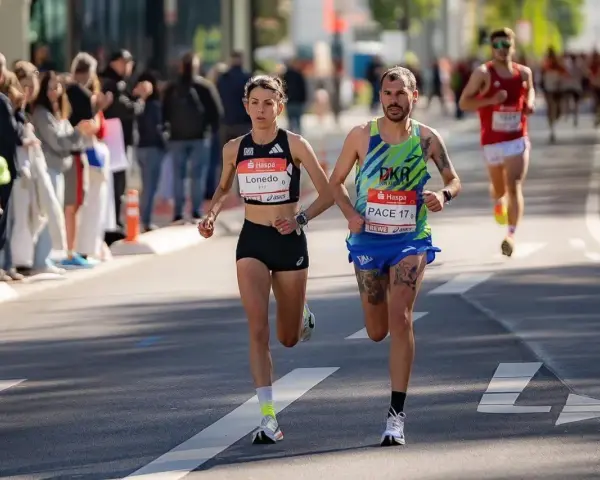 Atleti si preparano a una maratona, correndo fianco a fianco. La folla incita lungo il percorso.