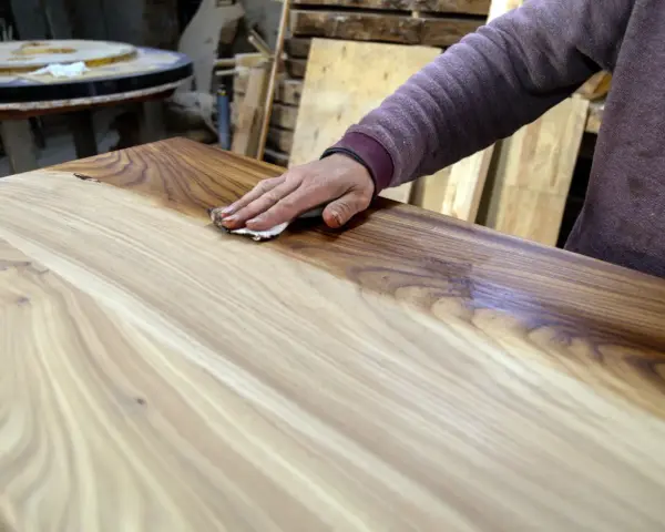 Mano che applica la finitura a cera dopo impregnante su un tavolo di legno grezzo, esaltandone le venature.