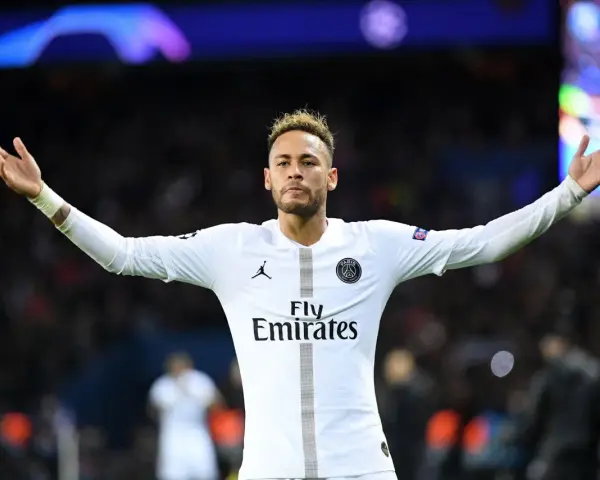 Neymar w białej koszulce PSG, z charakterystyczną neymar fryzurą, unosi ręce w geście radości na tle rozmazanych świateł stadionu.
