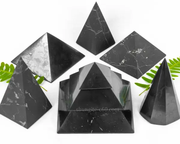 Seis pirámides de piedra shungita, algunas con vetas blancas, dispuestas sobre un fondo blanco con hojas verdes.
