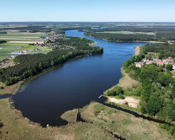 Malownicze jezioro Wojnowskie otoczone lasami i polami. W oddali widać zabudowania wsi, a na brzegu plażę z pomostem.
