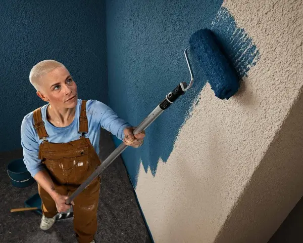 Frau mit Kurzhaarschnitt streicht mit blauem Roller eine Wand mit Rauputz.