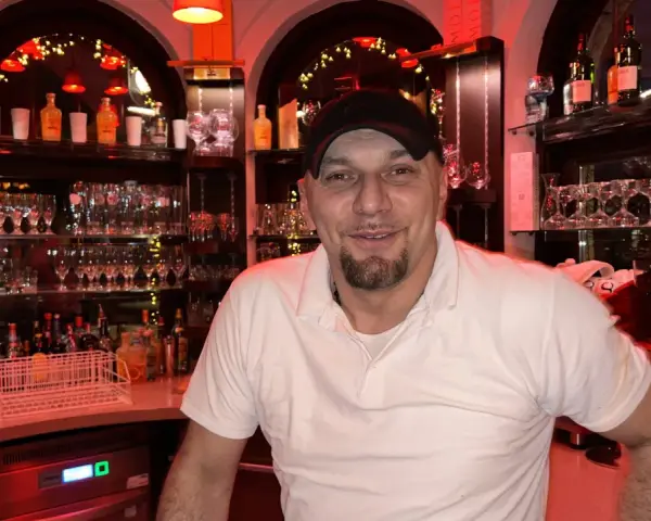 Mann mit Bart und Kappe hinter der Bar, umgeben von Gläsern und Flaschen. Er lächelt freundlich, bereit für die nächste Bestellung in "la gondola due".