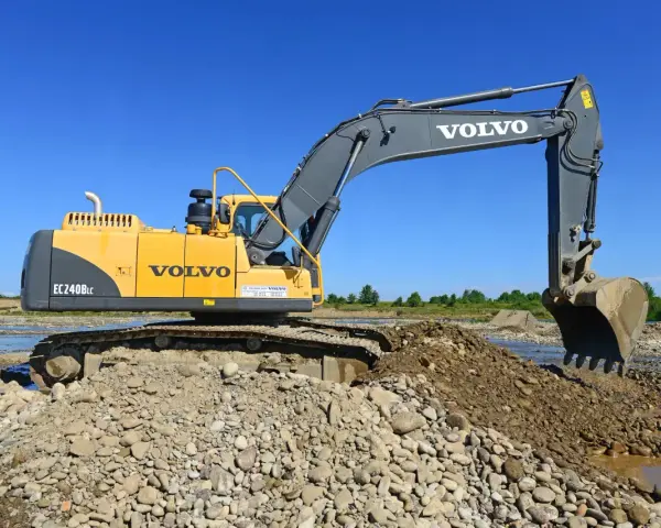 Żółta koparka Volvo EC240B LC pracuje na kamienistym brzegu rzeki. Zastanawiasz się, ile kosztuje godzina pracy koparki?