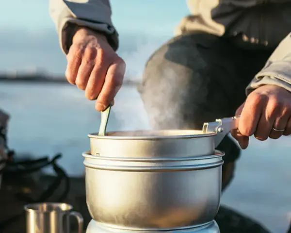 En person lagar mat utomhus, ångande kastruller och en mugg i metall antyder en lyxig matupplevelse i naturen.