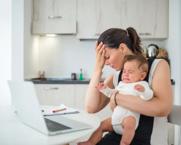 Zmęczona mama z płaczącym niemowlakiem na rękach, próbuje pracować przy laptopie. Czuje przebodźcowanie niemowlaka i własne.