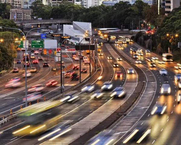 Traffico intenso su un'autostrada urbana al tramonto. Le luci dei veicoli creano scie luminose, con un **segnale 105** che indica la direzione.