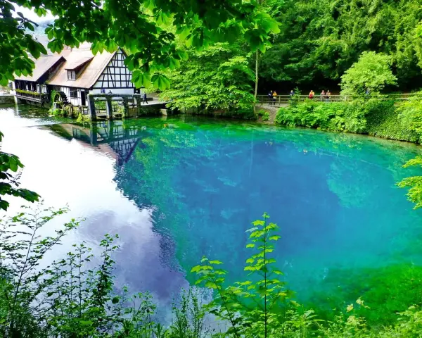 Der Blautopf See lädt zum Schwimmen ein. Ein historisches Fachwerkhaus mit Wasserrad spiegelt sich im kristallklaren, blauen Wasser.