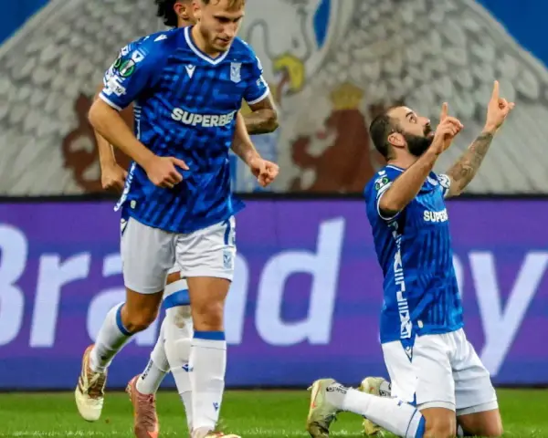 Piłkarz klęczy na boisku, celebrując bramkę. W tle biegnie kolega z drużyny. Wszyscy w niebieskich strojach. Czekamy, kiedy wraca ekstraklasa!