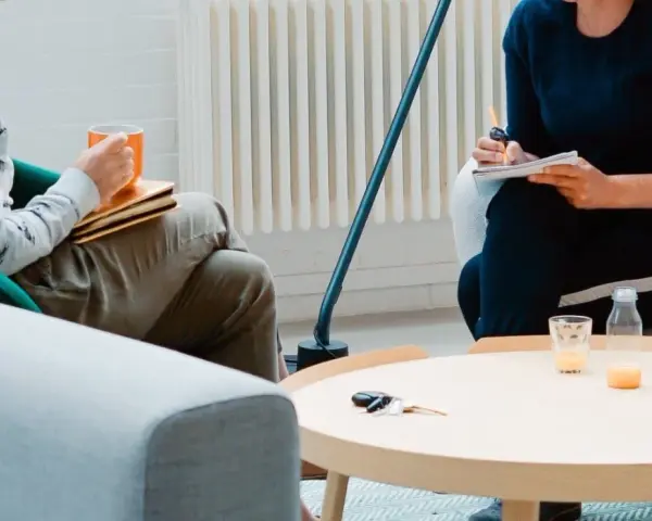 Dwie osoby siedzą w fotelach, jedna trzyma kubek, druga pisze w notatniku.