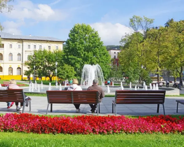 Park z fontanną i ludźmi siedzącymi na ławkach, w tle budynek.