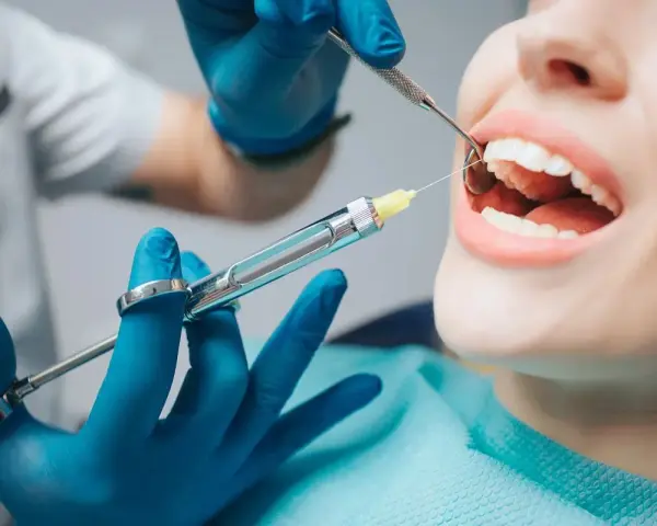 Dentysta podaje znieczulenie, by leki przeciwbólowe zadziałały przed zabiegiem. Pacjentka ma otwarte usta, a w nich widać narzędzia stomatologiczne.
