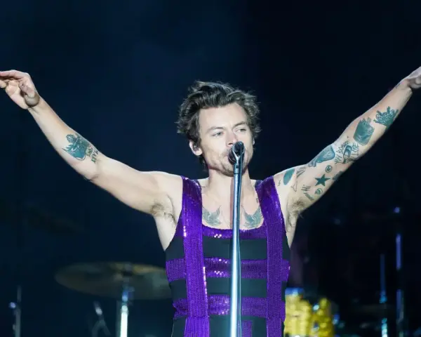 Harry Styles w błyszczącym fioletowym topie śpiewa na koncercie, unosząc ramiona. Jego ramiona pokryte są tatuażami.