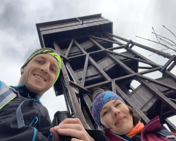 Para wędrowców uśmiecha się na tle drewnianej wieży widokowej. W tle majaczy **gór**ski krajobraz.