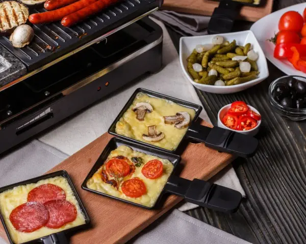 Raclette z pieczarkami i pomidorami, obok kiełbaski. Idealny ser do raclette rozpuszcza się w tych małych patelenkach.