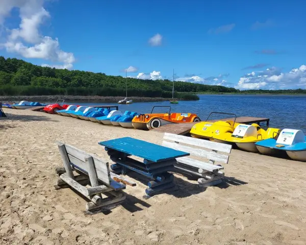 Łódki wodne czekają na plaży nad jeziorem Sarbsko. Drewniane ławki i stół zapraszają do odpoczynku.