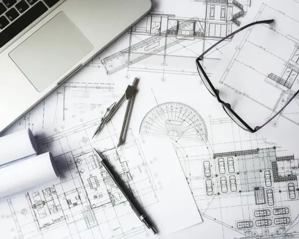 Na biurku architekta leżą plany budowlane, laptop, okulary i cyrkiel. Trwają prace nad przedmiarem robót.
