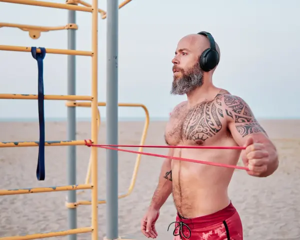 Man med tatueringar och skägg tränar med gummisnoddar vid ett utegym på stranden.