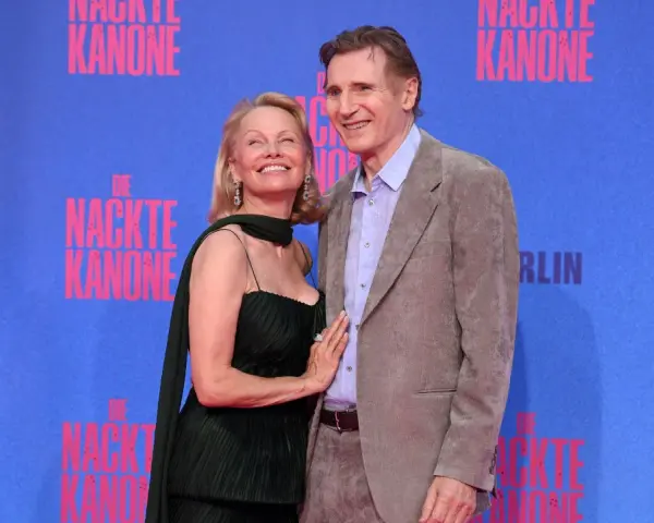 Liam Neeson e Pamela Anderson sorridono sul tappeto blu, con lo sfondo di "Die Nackte Kanone".