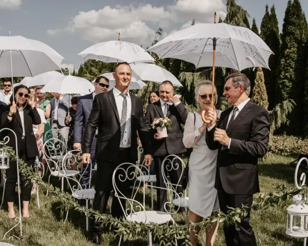 Wesele w kwietniu pod białymi parasolami. Goście w eleganckich strojach, wśród nich pan młody z bukiecikiem.