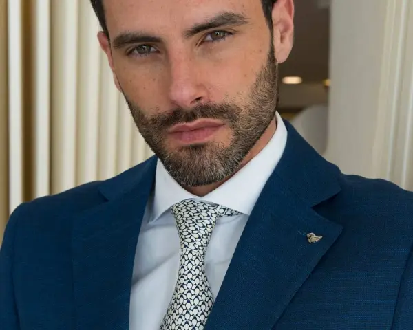 Uomo con abito blu, camicia bianca e cravatta con fantasia a pois bianchi e neri. Un tocco di stile con un pin a forma di ala.