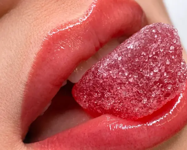 Labbra carnose e lucide, con un effetto volume dato da un tatuaggio labbra, assaporano una caramella rossa zuccherina.