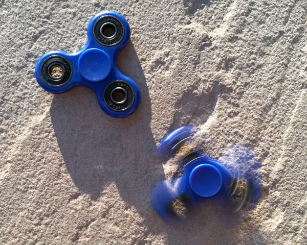 Dwa niebieskie fidget spinnery na piasku, jeden nieruchomy, drugi w ruchu, przypominają czasy, kiedy fidget spinnery były popularne.