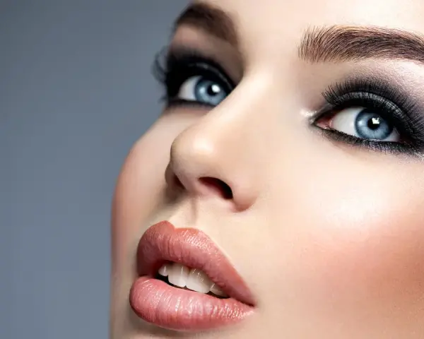 Niebieskie oczy z perfekcyjnym makijażem smokey eye, podkreślone długimi rzęsami i błyszczącymi ustami.