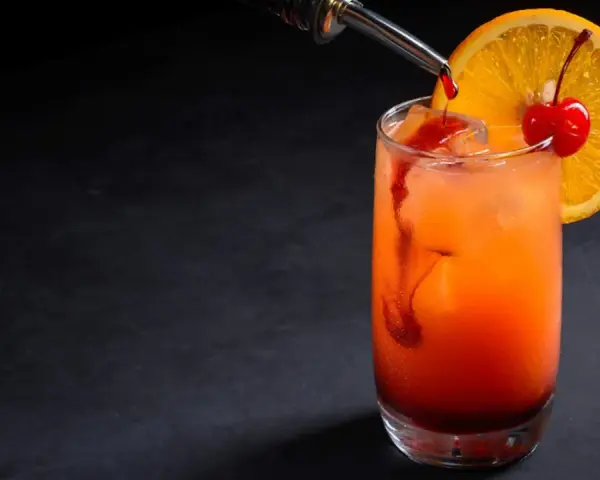 Kolorowy drink z syropem grenadina, ozdobiony plasterkiem pomarańczy i wisienką.