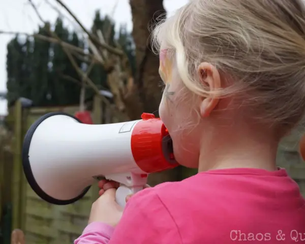 Ein Mädchen mit blonden Zöpfen hält ein Megafon und schreit hinein. Vielleicht fragt sie sich, warum sind Kinder so laut?