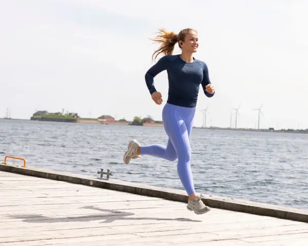 En kvinna springer glatt på en träbrygga vid havet, med vindkraftverk i bakgrunden. Hon verkar njuta av att springa 1 km varje dag.