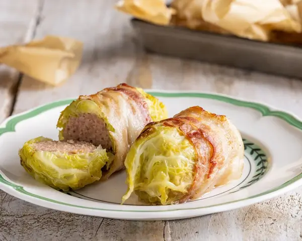 Deliziosi involtini di verza con carne, avvolti in pancetta croccante, serviti su un piatto bianco con bordo verde.