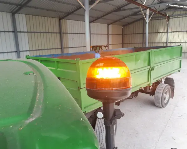 Pomarańczowa lampa ostrzegawcza na wysięgniku, przy zielonym ciągniku i przyczepie.