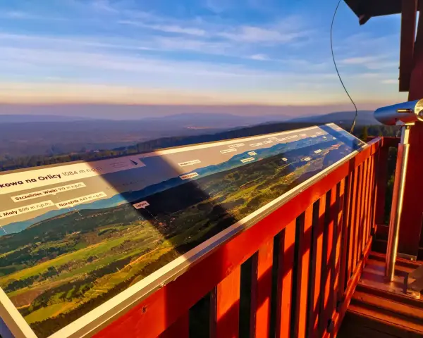 Widok z wieży widokowej na Orlicy. Na pierwszym planie panorama z opisem szczytów i dolin. W oddali łagodne wzgórza o zachodzie słońca.