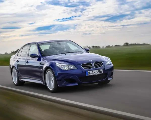 Niebieskie BMW M5 E60, ikona designu Chrisa Bangle'a, pędzi po drodze wśród zielonych pól i błękitnego nieba.