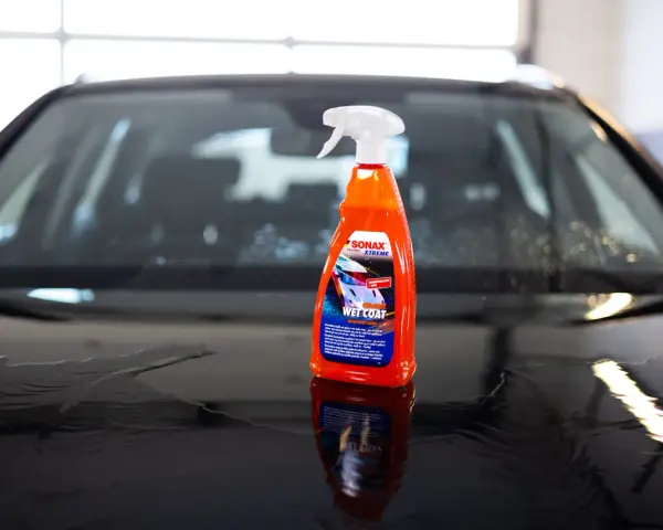 En flaska SONAX Xtreme Wet Coat står på en våt, mörk bilhuv.