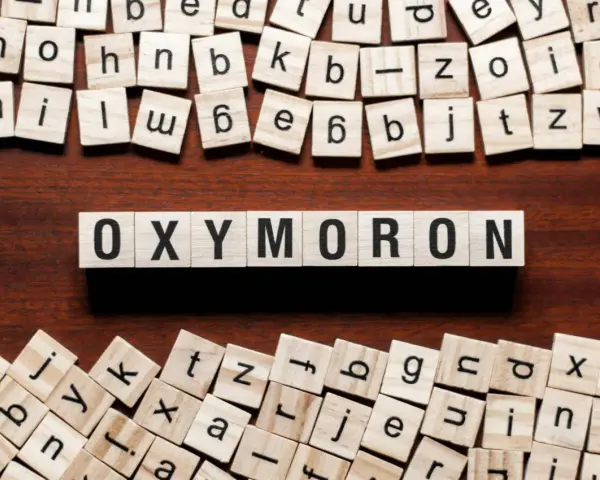 Holzbuchstaben bilden das Wort "OXYMORON". Sie sind umgeben von weiteren Buchstabenkacheln, die an Spiele wie Scrabble erinnern und Beispiele für Oxymoron-Begriffe darstellen könnten.