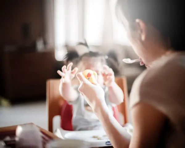 Niania karmi dziecko, które trzyma zabawkę. Zastanawiasz się, ile kosztuje godzina niani do dziecka?