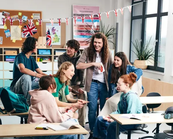 Uśmiechnięty nauczyciel z długimi włosami rozdaje karty uczniom w klasie. Wszyscy wyglądają na zaangażowanych w grę.