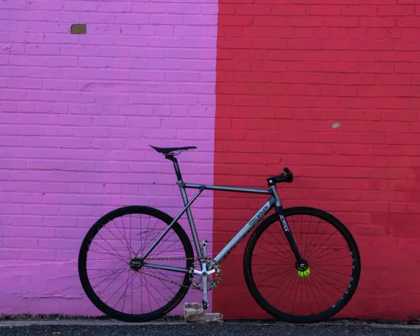 A sleek, fixed gear bike with "MASH" branding leans against a vibrant pink and red brick wall.